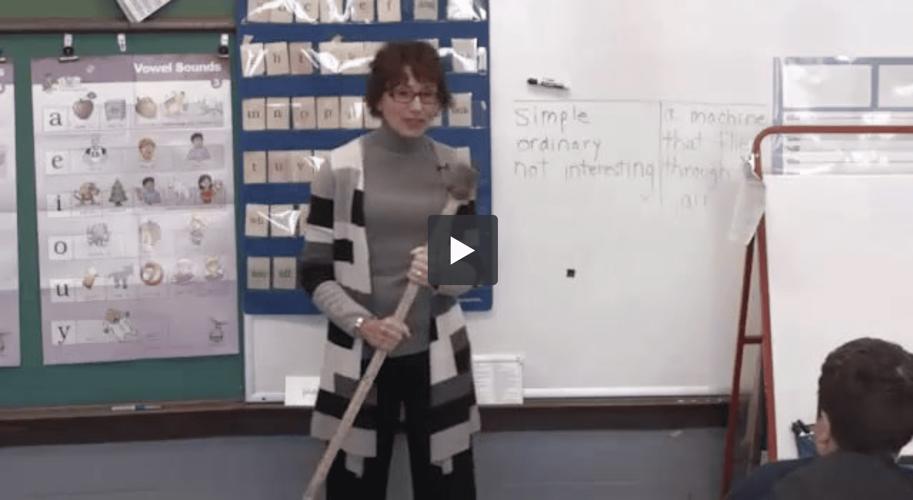 A teacher stands holding a large pointer stick in front of a whiteboard in a classroom. The whiteboard has text that reads Simple, ordinary, not interesting. Classroom posters and charts related to vowel sounds are visible on the wall beside her. Students are sitting at desks.